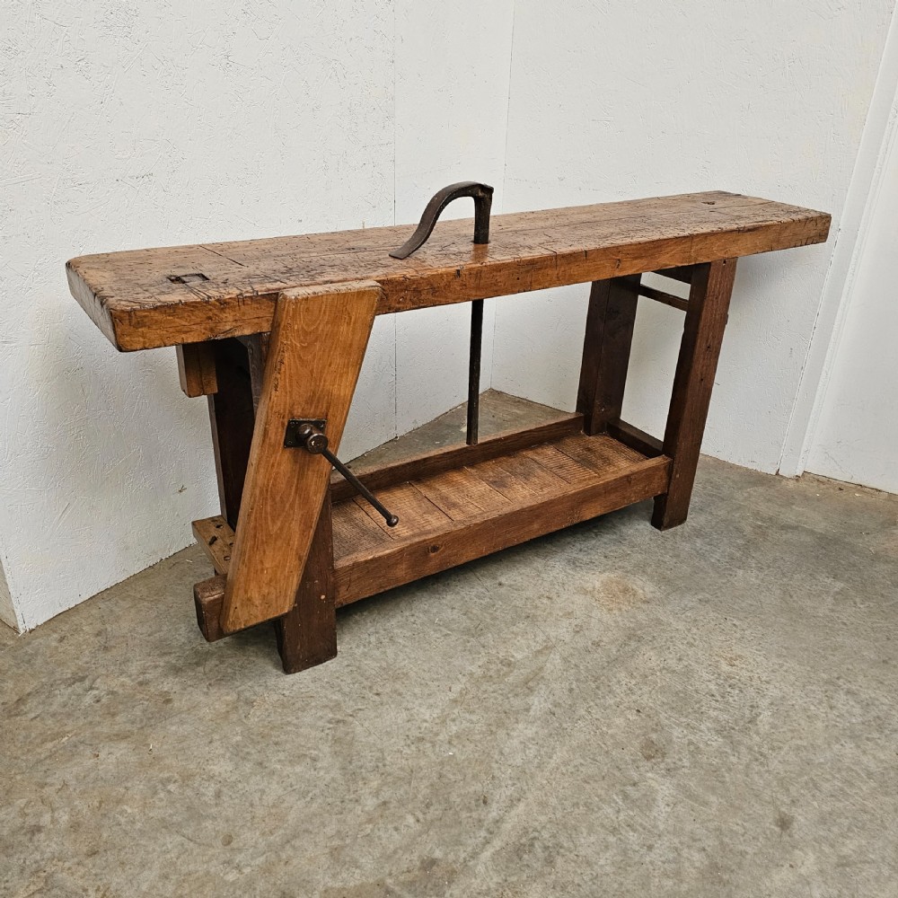 A rustic wooden French artisan workbench featuring a thick timber top, integrated wooden leg vise, iron holdfast tool, and a low slatted storage shelf.