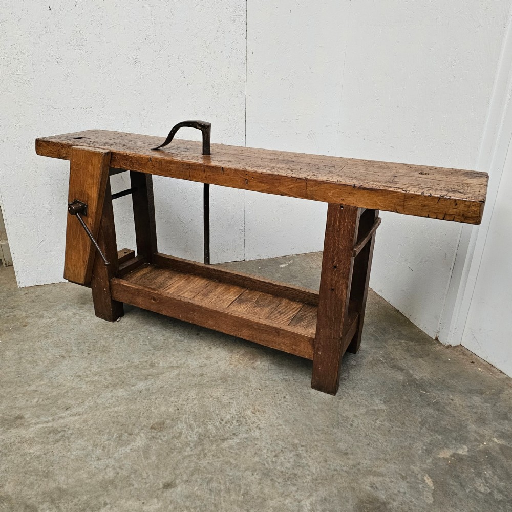 A rustic wooden French artisan workbench featuring a thick timber top, integrated wooden leg vise, iron holdfast tool, and a low slatted storage shelf. View II