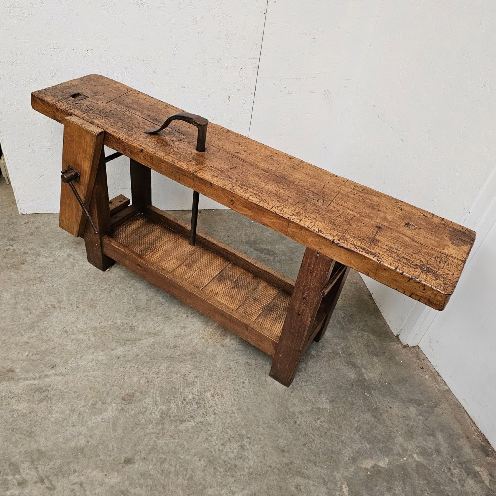 A rustic wooden French artisan workbench featuring a thick timber top, integrated wooden leg vise, iron holdfast tool, and a low slatted storage shelf. View III