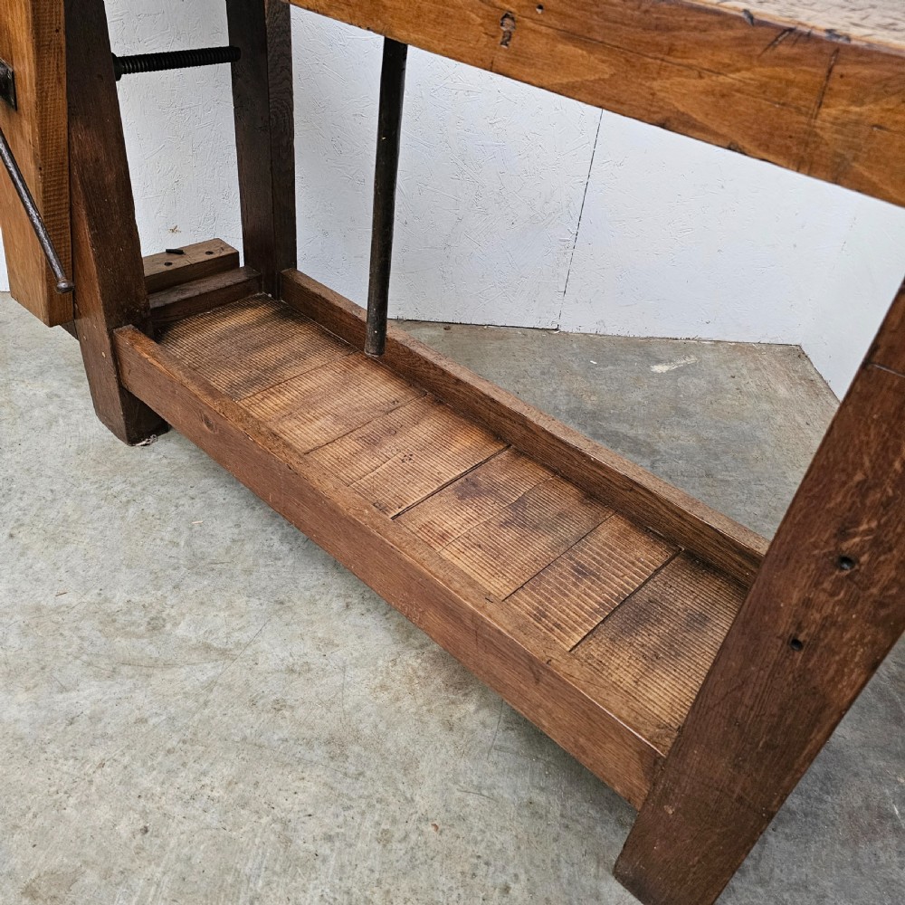 A rustic wooden French artisan workbench featuring a thick timber top, integrated wooden leg vise, iron holdfast tool, and a low slatted storage shelf. View IV