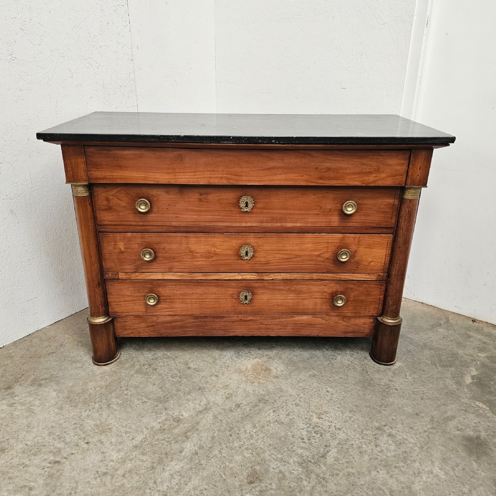 A French Empire warm brown timber chest of drawers featuring four drawers, flanking columns with brass mounts, brass knobs, and a black marble top.