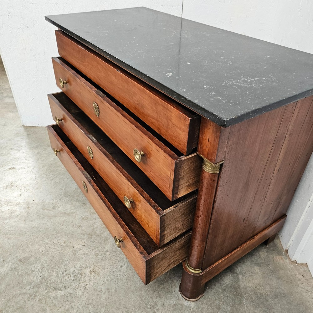 A French Empire warm brown timber chest of drawers featuring four drawers, flanking columns with brass mounts, brass knobs, and a black marble top. View II