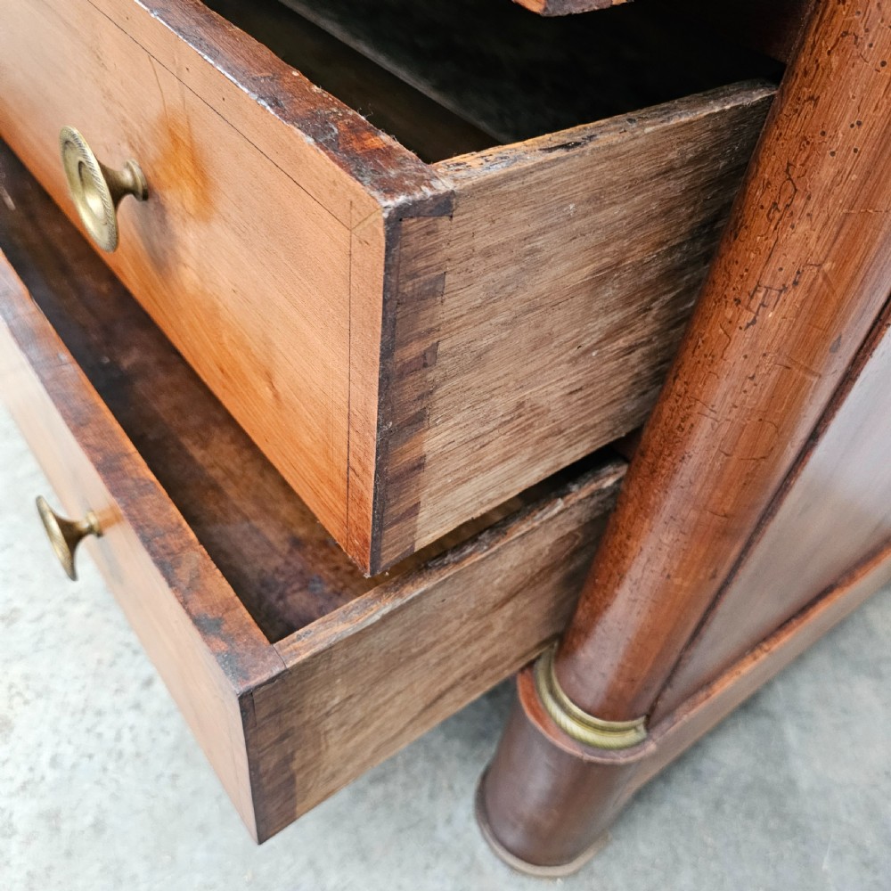 A French Empire warm brown timber chest of drawers featuring four drawers, flanking columns with brass mounts, brass knobs, and a black marble top. View V
