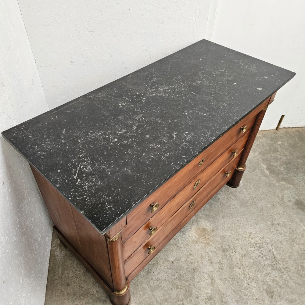 A French Empire warm brown timber chest of drawers featuring four drawers, flanking columns with brass mounts, brass knobs, and a black marble top. View VI
