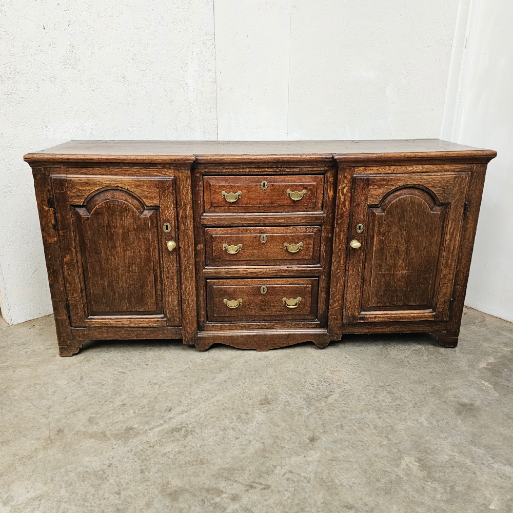 A Georgian oak breakfront dresser base featuring a central stack of three drawers flanked by two arched panel cupboard doors with brass handles.