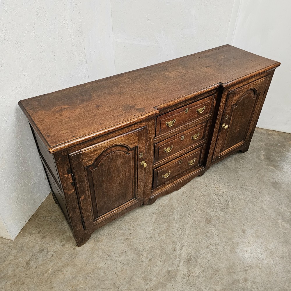 A Georgian oak breakfront dresser base featuring a central stack of three drawers flanked by two arched panel cupboard doors with brass handles. View II