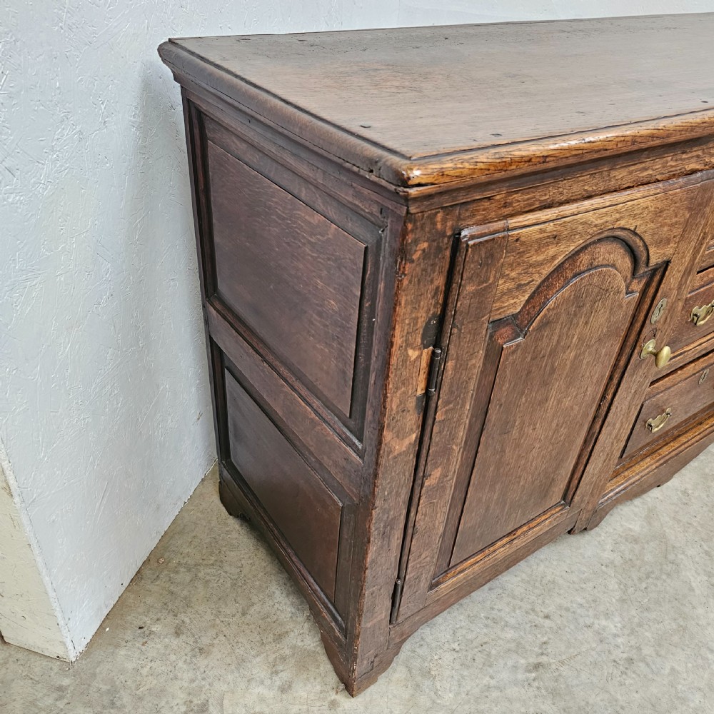 A Georgian oak breakfront dresser base featuring a central stack of three drawers flanked by two arched panel cupboard doors with brass handles. View III