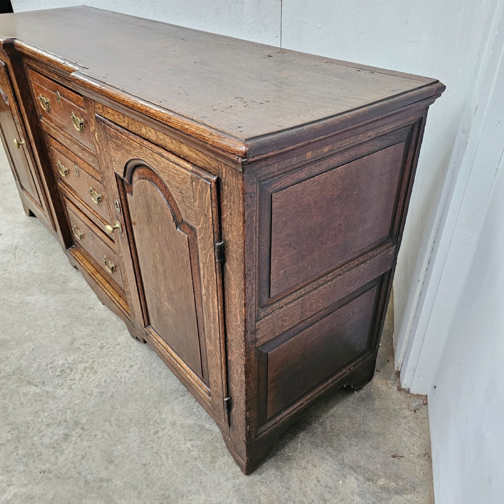 A Georgian oak breakfront dresser base featuring a central stack of three drawers flanked by two arched panel cupboard doors with brass handles. View V