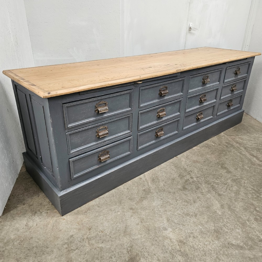A long French shop counter featuring twelve drawers painted in dark grey with metal cup handles and a natural scrubbed pine top surface.