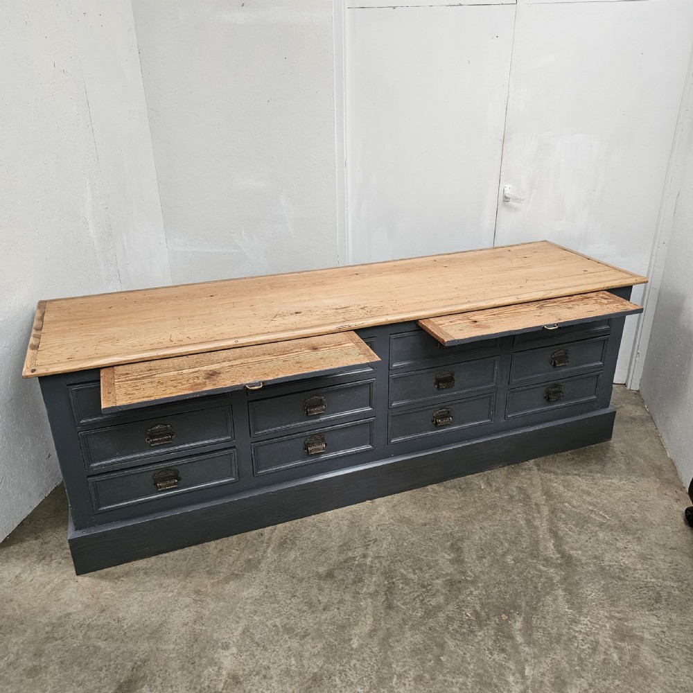 A long French shop counter featuring twelve drawers painted in dark grey with metal cup handles and a natural scrubbed pine top surface. View III