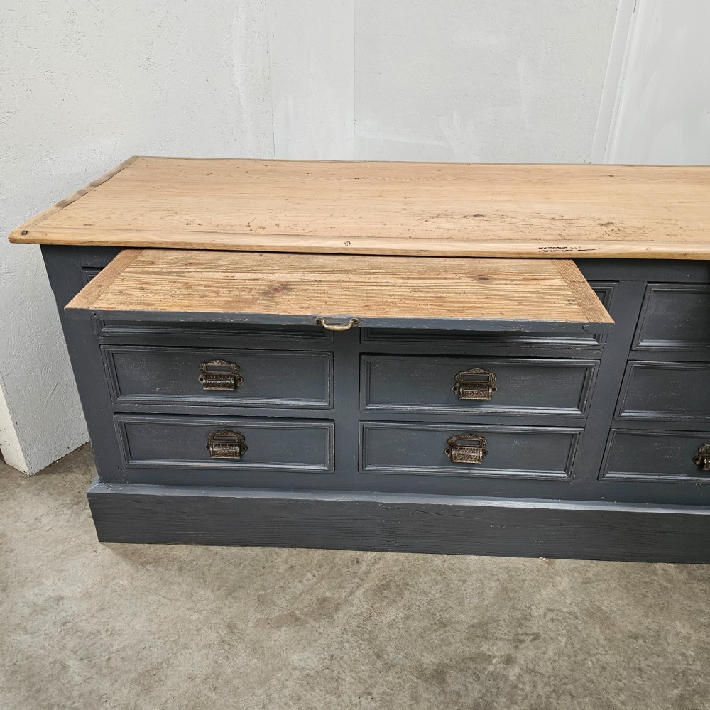 A long French shop counter featuring twelve drawers painted in dark grey with metal cup handles and a natural scrubbed pine top surface. View IV