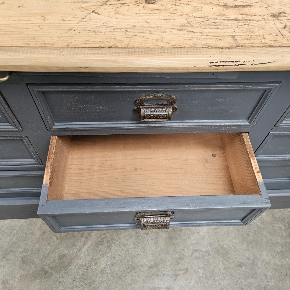 A long French shop counter featuring twelve drawers painted in dark grey with metal cup handles and a natural scrubbed pine top surface. View V