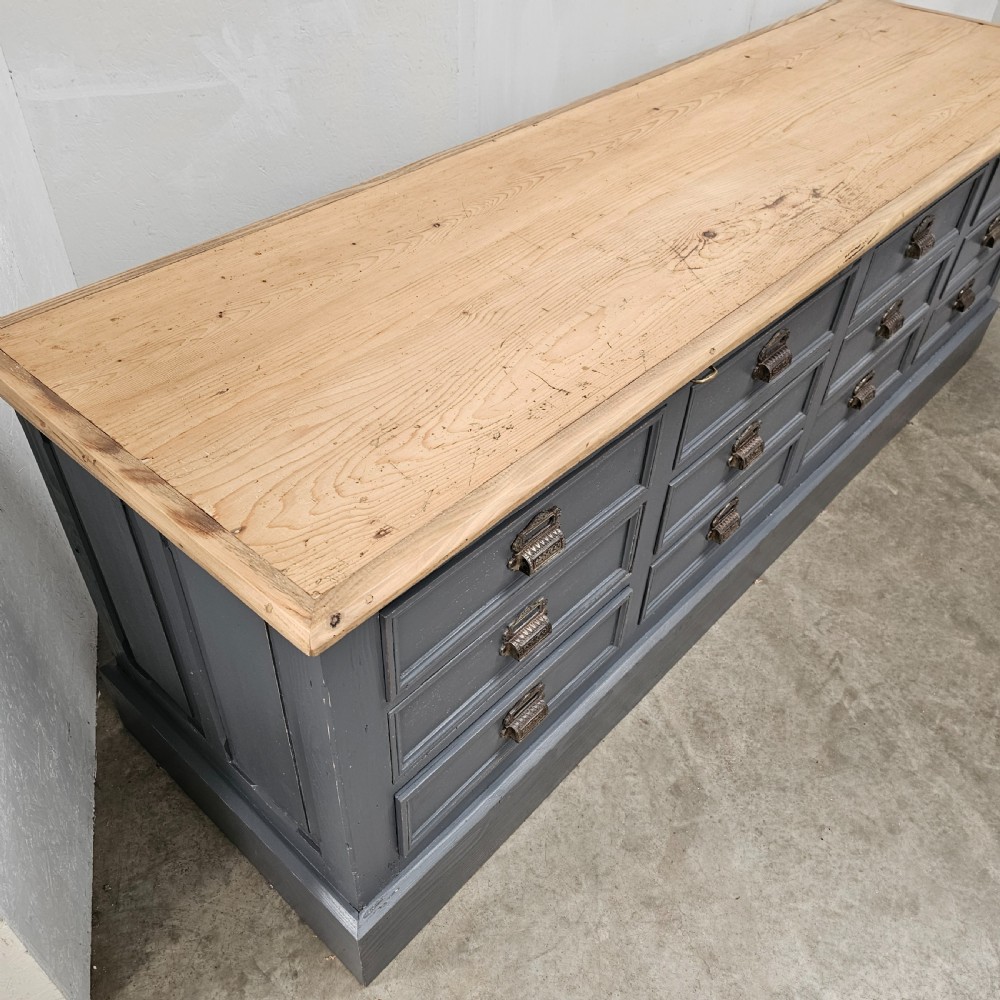 A long French shop counter featuring twelve drawers painted in dark grey with metal cup handles and a natural scrubbed pine top surface. View VII