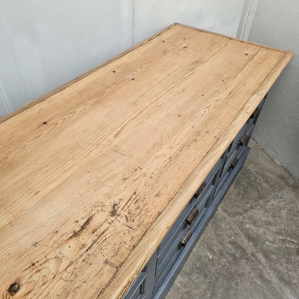 A long French shop counter featuring twelve drawers painted in dark grey with metal cup handles and a natural scrubbed pine top surface. View VIII