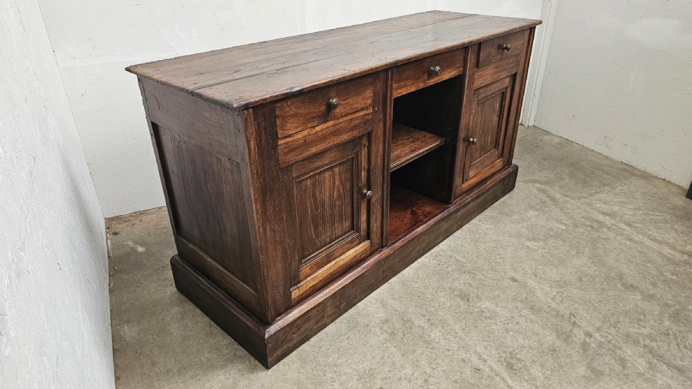 A 19th-century French dark chestnut dresser base featuring three top drawers above two panelled cupboard doors and a central open shelving section, set on a solid plinth base. View II