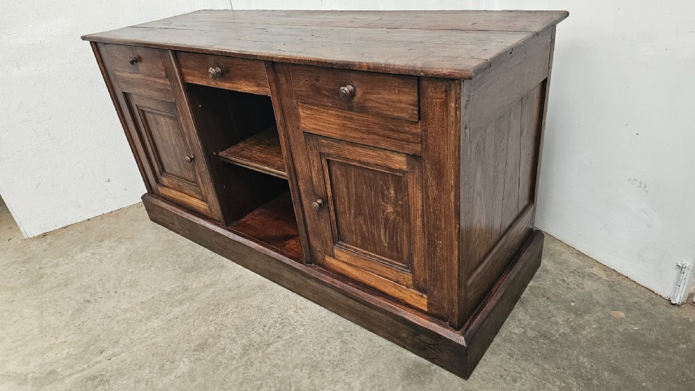 A 19th-century French dark chestnut dresser base featuring three top drawers above two panelled cupboard doors and a central open shelving section, set on a solid plinth base. View III