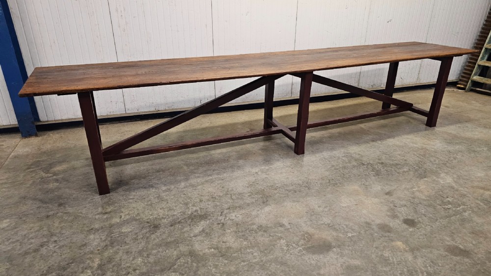 An exceptionally long antique pine harvest table featuring a plank wood top supported by a dark stained frame with cross-bracing.