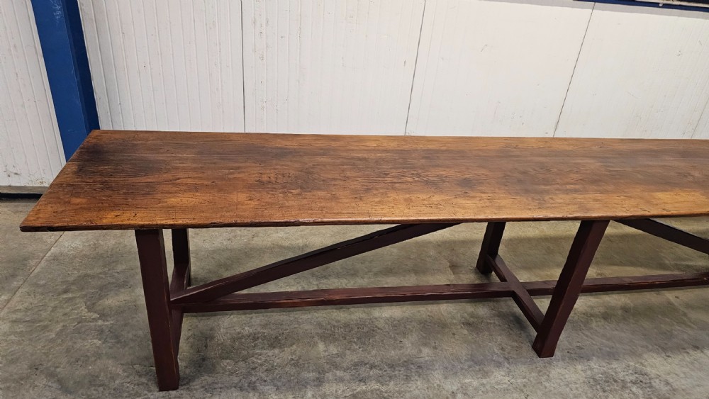 An exceptionally long antique pine harvest table featuring a plank wood top supported by a dark stained frame with cross-bracing. View V