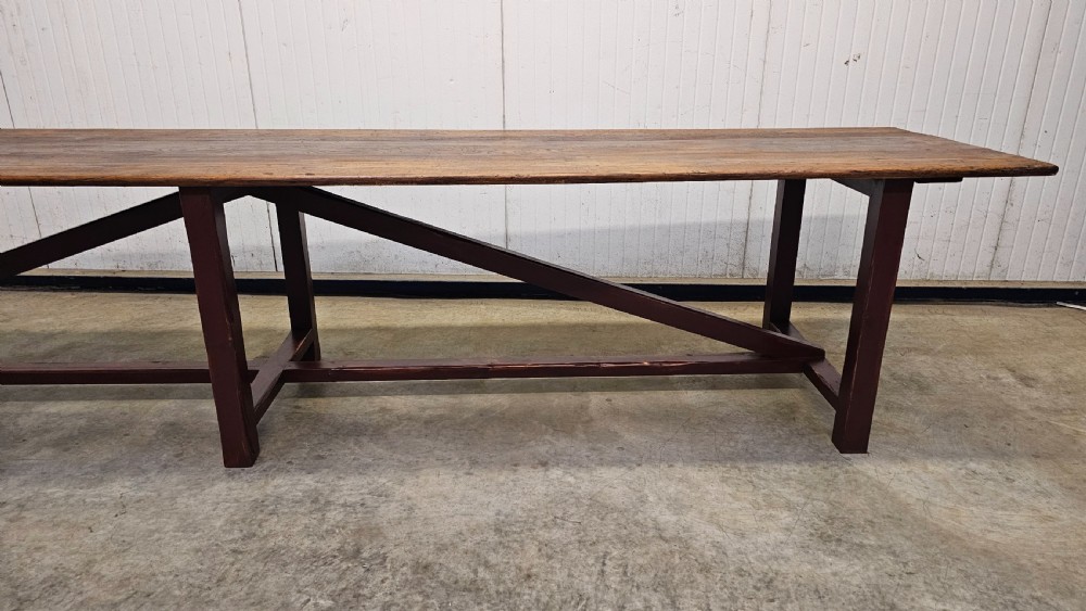 An exceptionally long antique pine harvest table featuring a plank wood top supported by a dark stained frame with cross-bracing. View VI