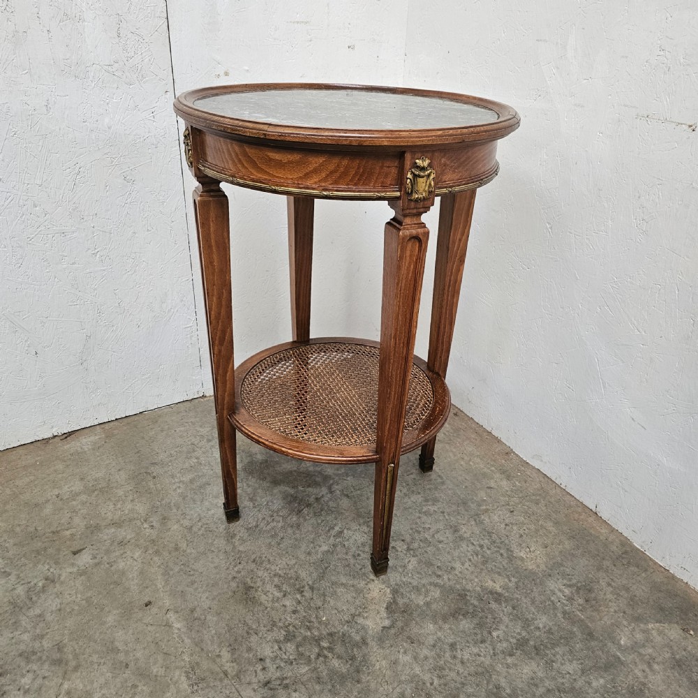 A French Empire style round wooden gueridon table featuring an inset dark marble top, brass mounts, tapering legs, and a cane lower shelf.