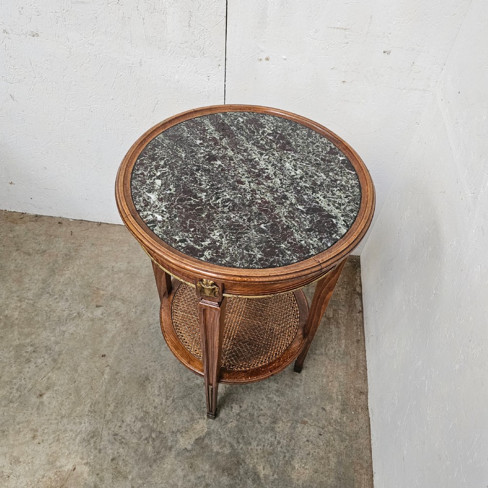 A French Empire style round wooden gueridon table featuring an inset dark marble top, brass mounts, tapering legs, and a cane lower shelf. View II