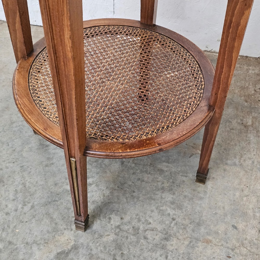 A French Empire style round wooden gueridon table featuring an inset dark marble top, brass mounts, tapering legs, and a cane lower shelf. View III