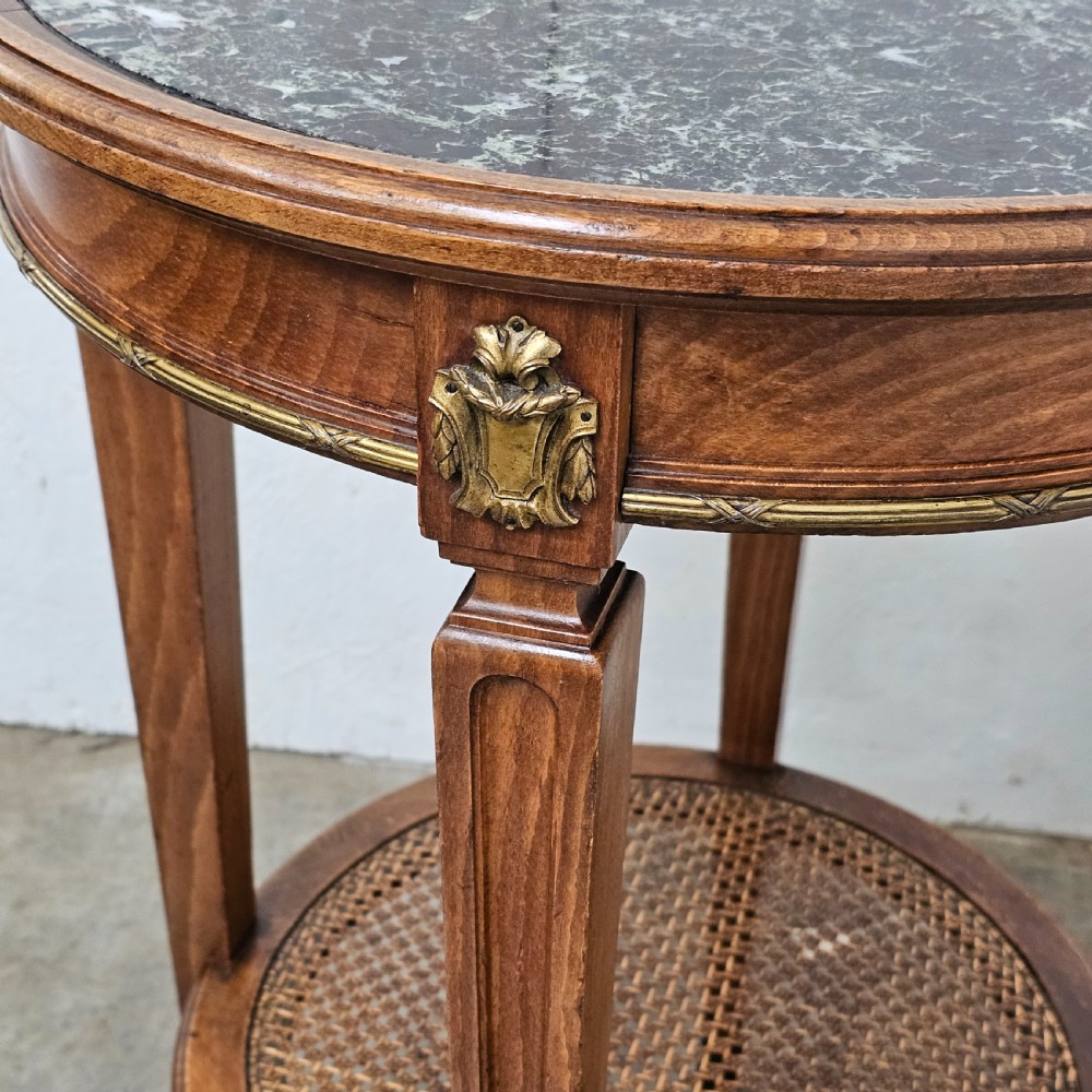 A French Empire style round wooden gueridon table featuring an inset dark marble top, brass mounts, tapering legs, and a cane lower shelf. View IV