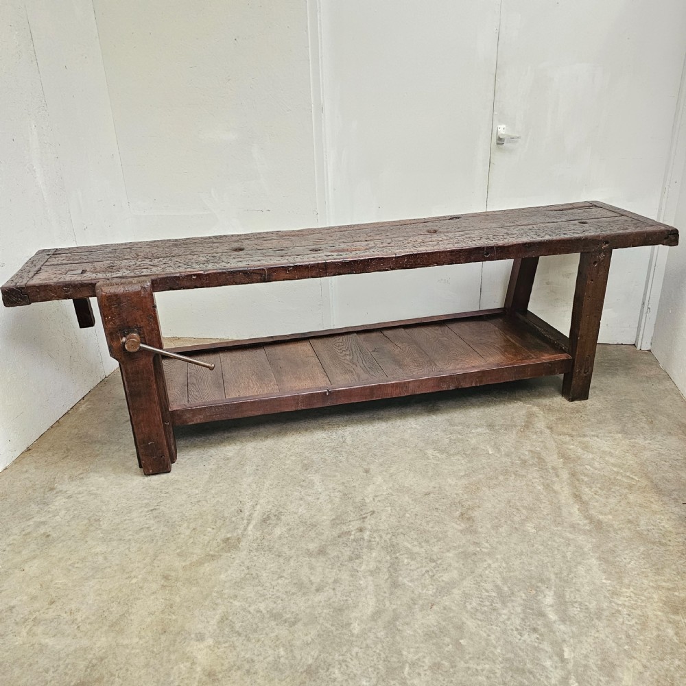 A long, dark oak antique workbench featuring an integrated wooden screw vice, a distressed top surface, and a full-length lower timber shelf.