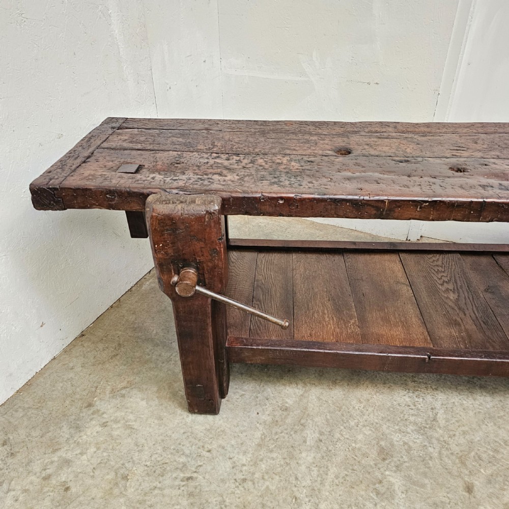 A long, dark oak antique workbench featuring an integrated wooden screw vice, a distressed top surface, and a full-length lower timber shelf. View II