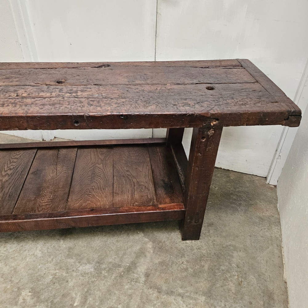 A long, dark oak antique workbench featuring an integrated wooden screw vice, a distressed top surface, and a full-length lower timber shelf. View III