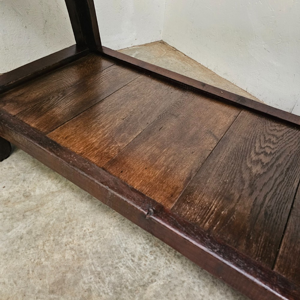 A long, dark oak antique workbench featuring an integrated wooden screw vice, a distressed top surface, and a full-length lower timber shelf. View V