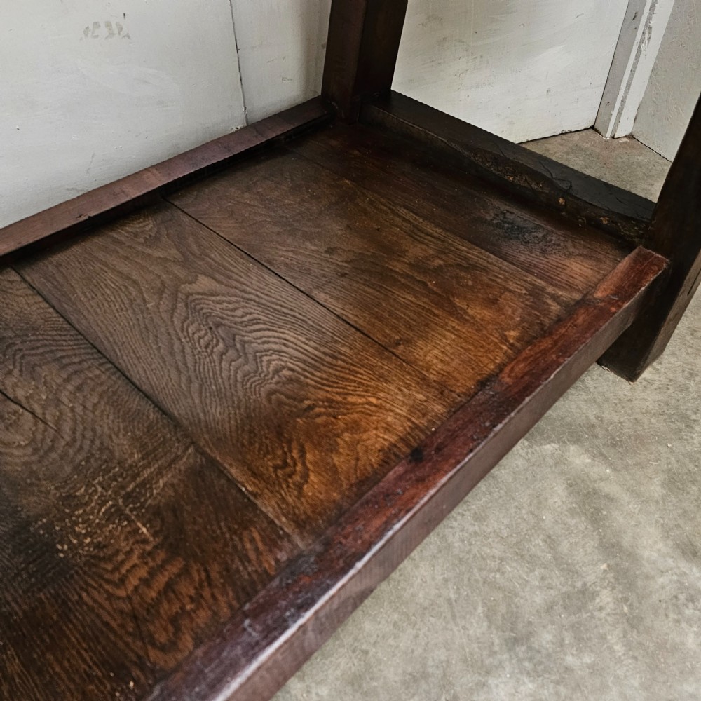 A long, dark oak antique workbench featuring an integrated wooden screw vice, a distressed top surface, and a full-length lower timber shelf. View VI