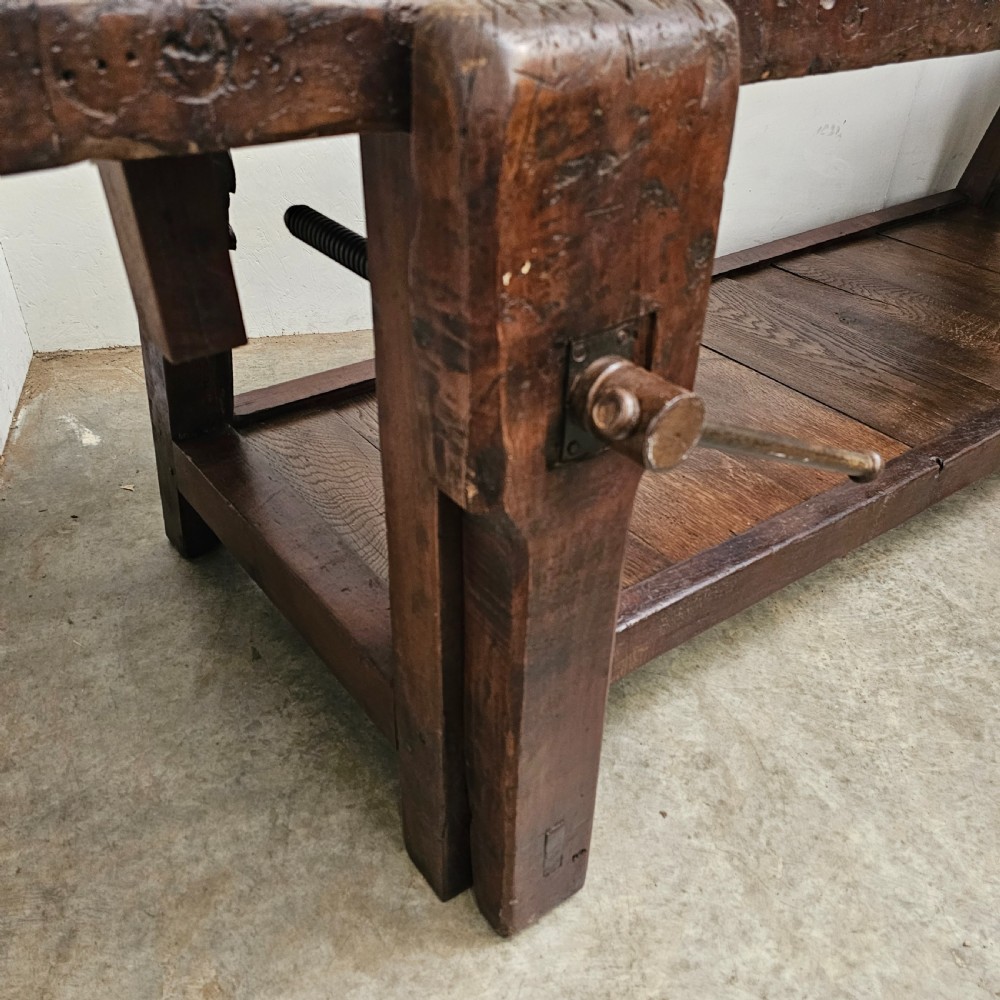 A long, dark oak antique workbench featuring an integrated wooden screw vice, a distressed top surface, and a full-length lower timber shelf. View VII