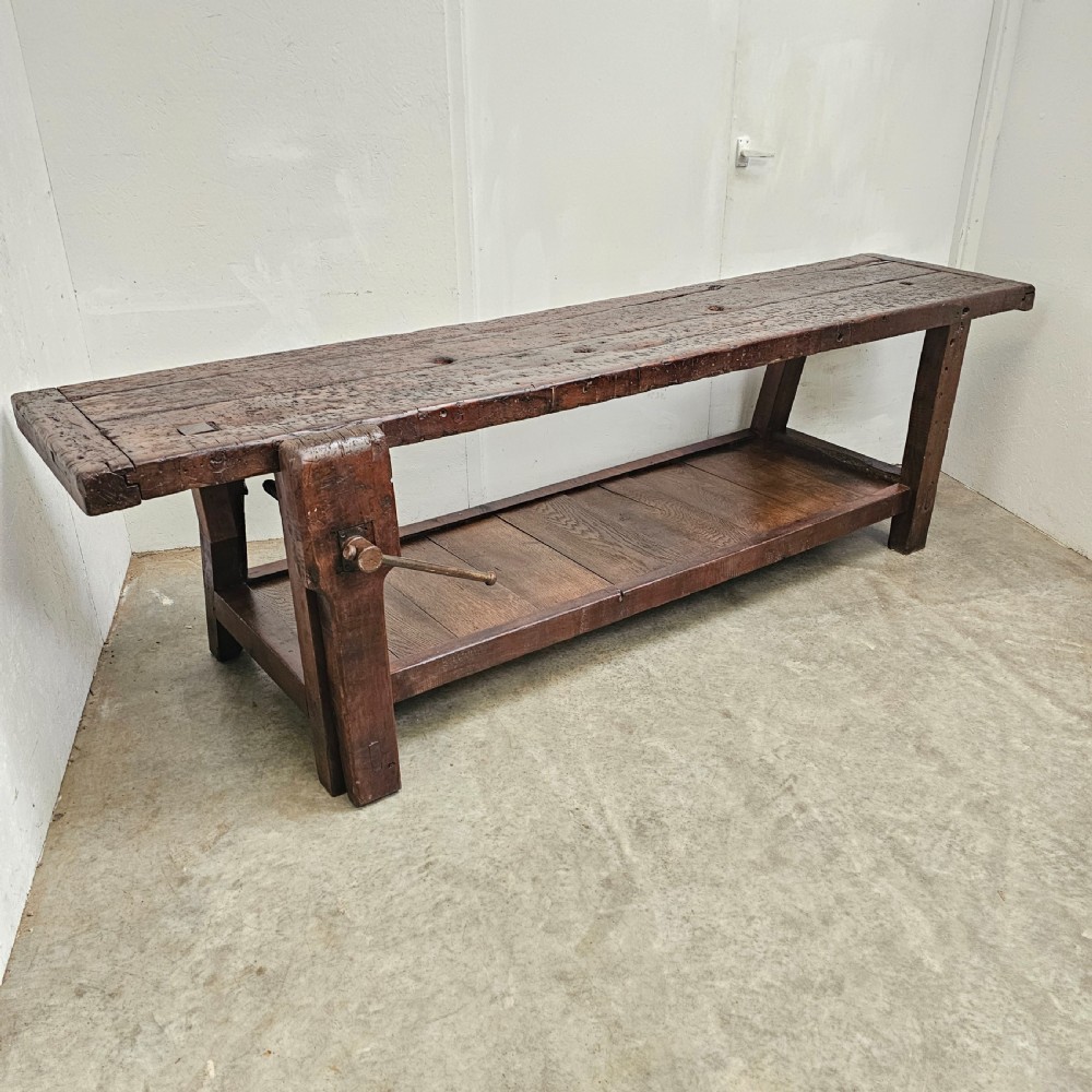 A long, dark oak antique workbench featuring an integrated wooden screw vice, a distressed top surface, and a full-length lower timber shelf. View VIII