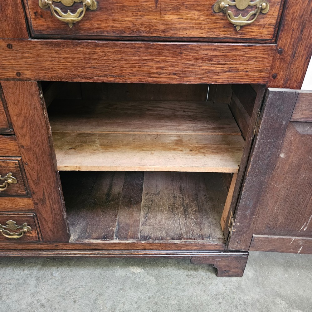 A 19th-century English oak dresser base featuring three upper drawers, three central drawers, two panelled cupboard doors, and brass swan-neck handles. View V