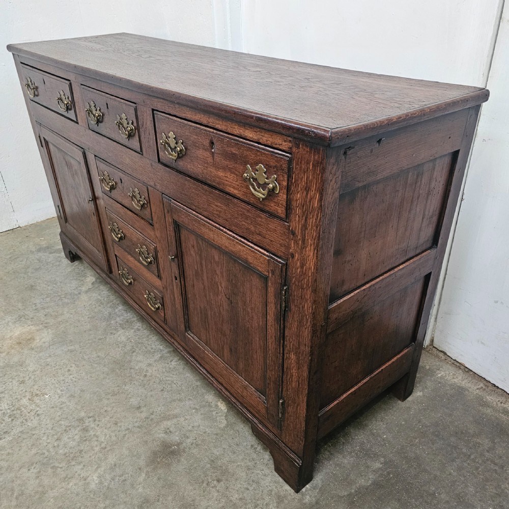 A 19th-century English oak dresser base featuring three upper drawers, three central drawers, two panelled cupboard doors, and brass swan-neck handles. View VI
