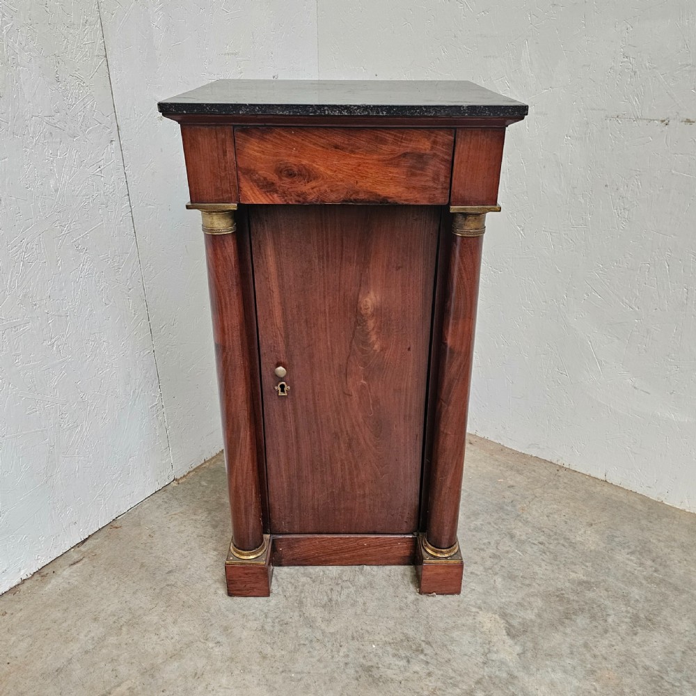 A French Empire style mahogany bedside cabinet featuring a dark marble top, single drawer, cupboard door, and flanking column supports with brass capitals.