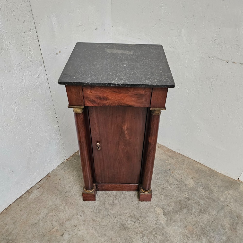 A French Empire style mahogany bedside cabinet featuring a dark marble top, single drawer, cupboard door, and flanking column supports with brass capitals. View II