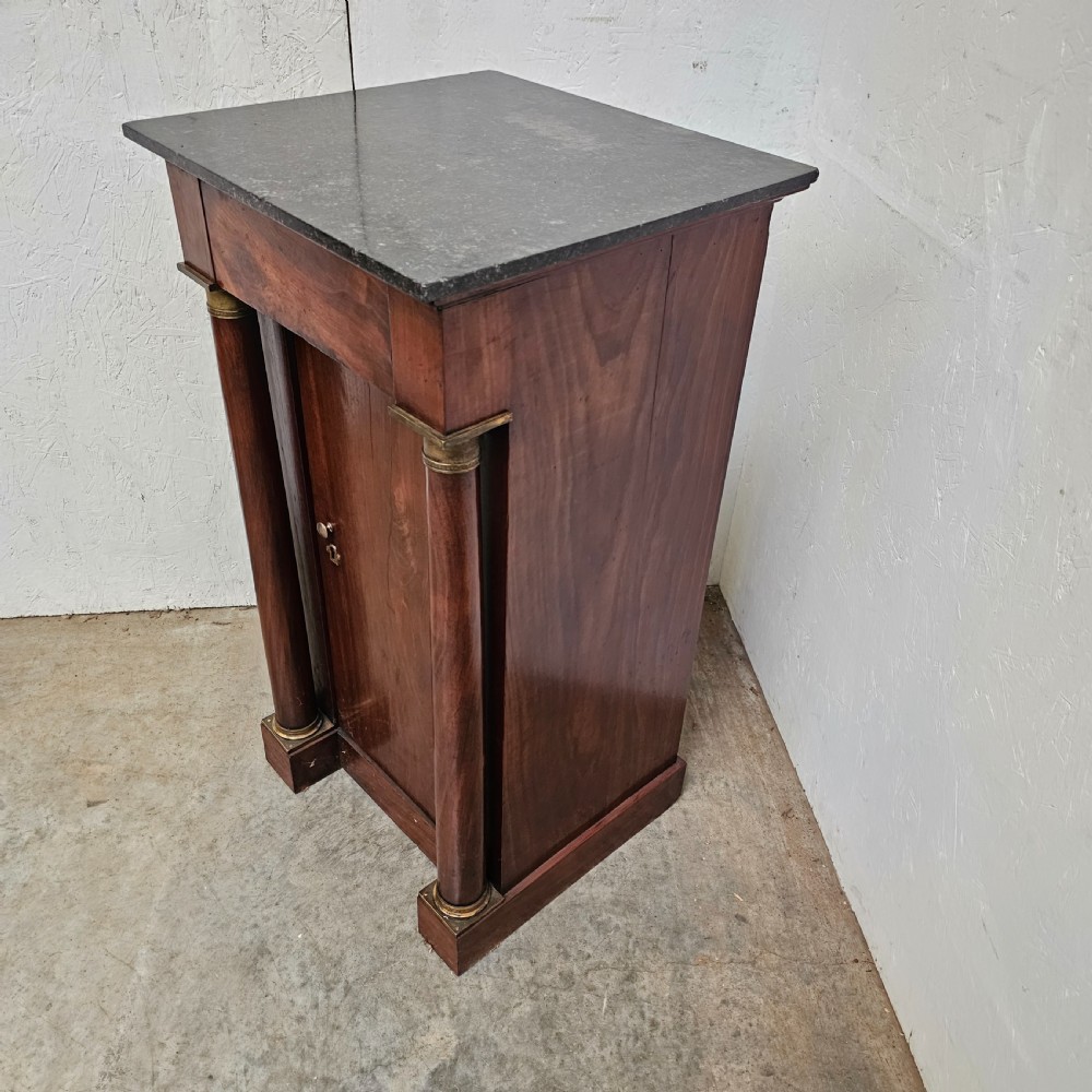 A French Empire style mahogany bedside cabinet featuring a dark marble top, single drawer, cupboard door, and flanking column supports with brass capitals. View III
