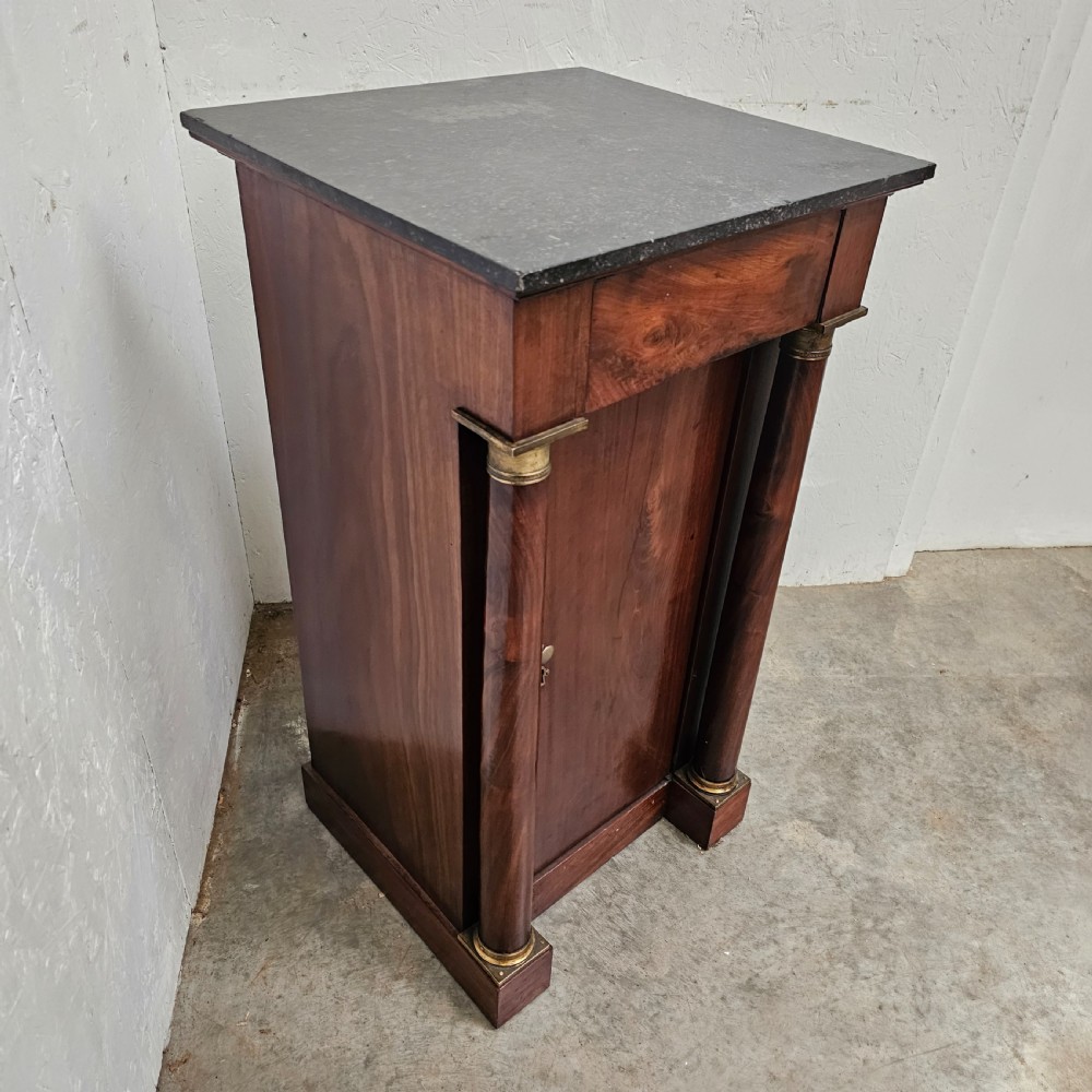 A French Empire style mahogany bedside cabinet featuring a dark marble top, single drawer, cupboard door, and flanking column supports with brass capitals. View V
