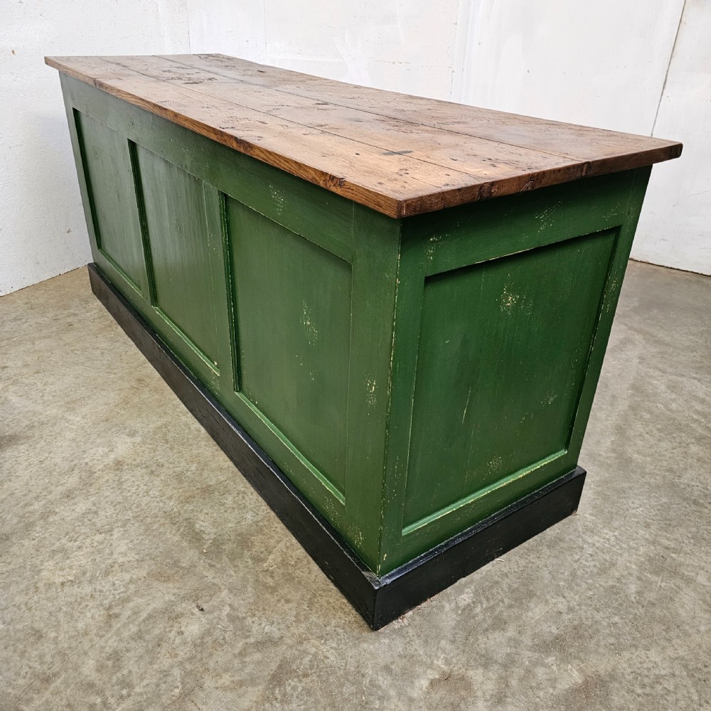 A long rustic French shop counter featuring a natural oak top, three recessed green painted front panels, and a black base plinth. View II