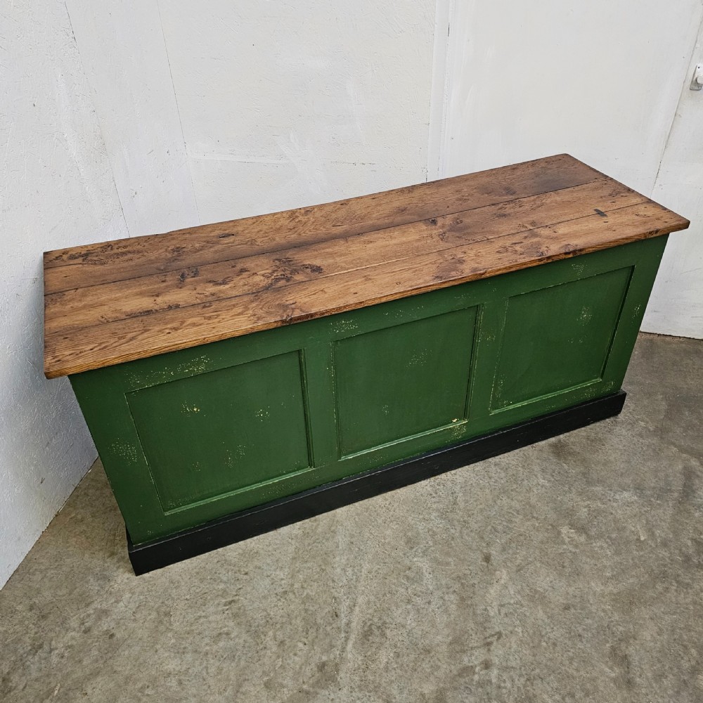 A long rustic French shop counter featuring a natural oak top, three recessed green painted front panels, and a black base plinth. View V