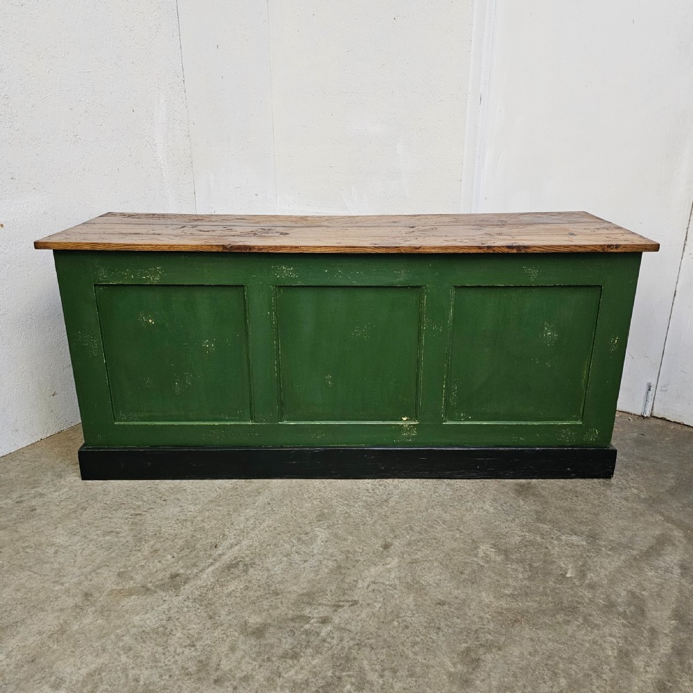A long rustic French shop counter featuring a natural oak top, three recessed green painted front panels, and a black base plinth.