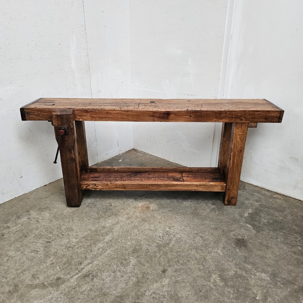 A rustic French wooden workbench featuring a thick plank top, lower shelf, and integrated vice on the left leg.