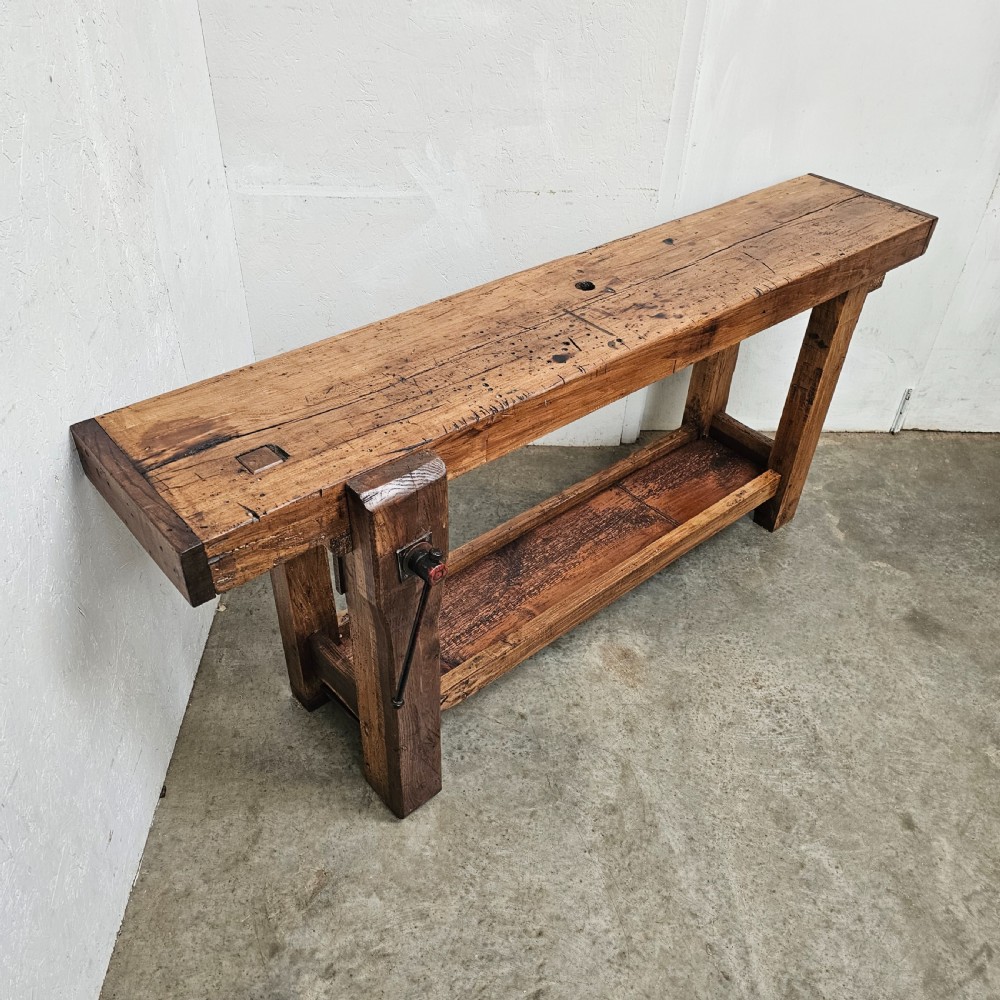 A rustic French wooden workbench featuring a thick plank top, lower shelf, and integrated vice on the left leg. View VI