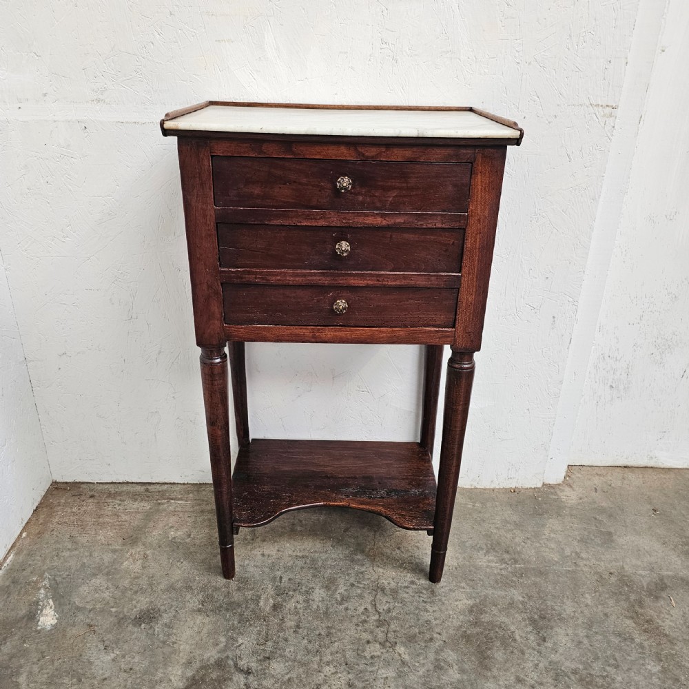 An antique French dark walnut bedside chest featuring an inset white marble top, three stacked drawers with metal rosette knobs, turned legs, and a lower undertier shelf.