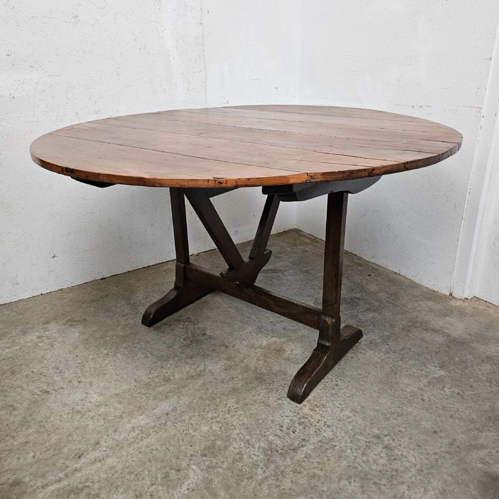 A large circular cherry wood plank table top resting on an oak trestle base in a tilted vertical orientation. View II
