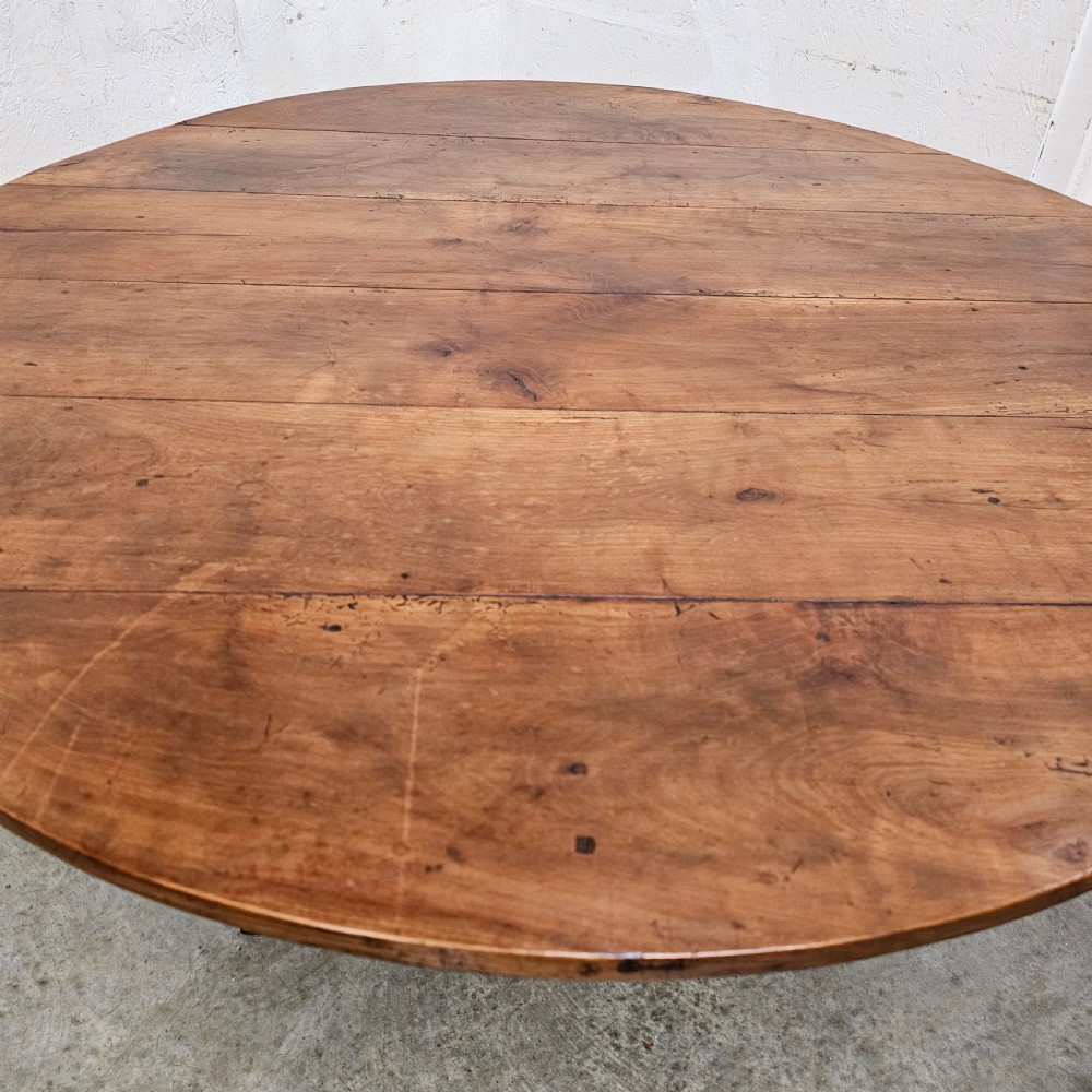 A large circular cherry wood plank table top resting on an oak trestle base in a tilted vertical orientation. View III