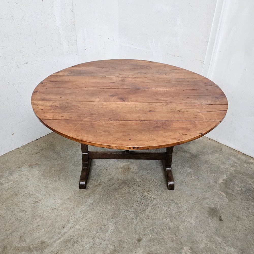 A large circular cherry wood plank table top resting on an oak trestle base in a tilted vertical orientation. View IV