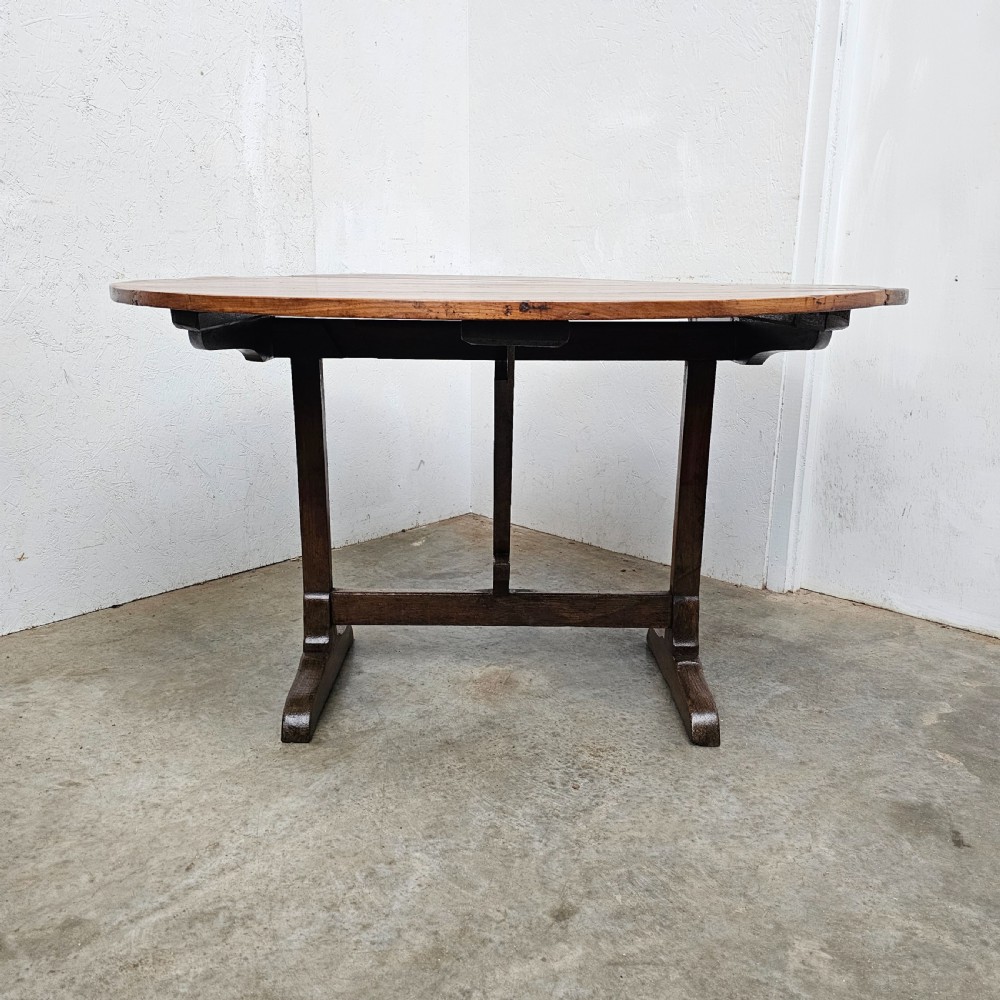 A large circular cherry wood plank table top resting on an oak trestle base in a tilted vertical orientation. View V